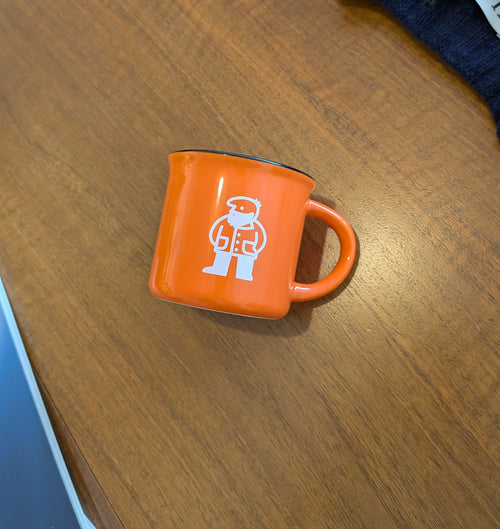 An orange enamel mug with a white astronaut graphic, resting on a wooden table next to a blue book and some folded clothing.