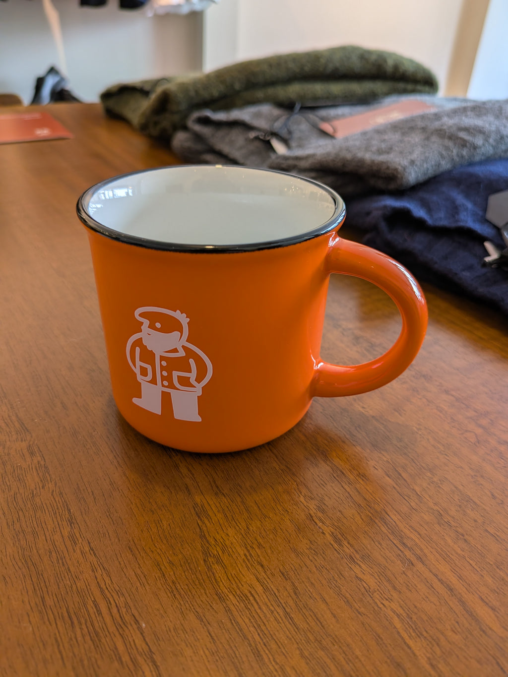 An orange enamel mug with a white graphic of a person wearing a cap and holding a phone, sitting on a wooden table with folded clothes in the background.