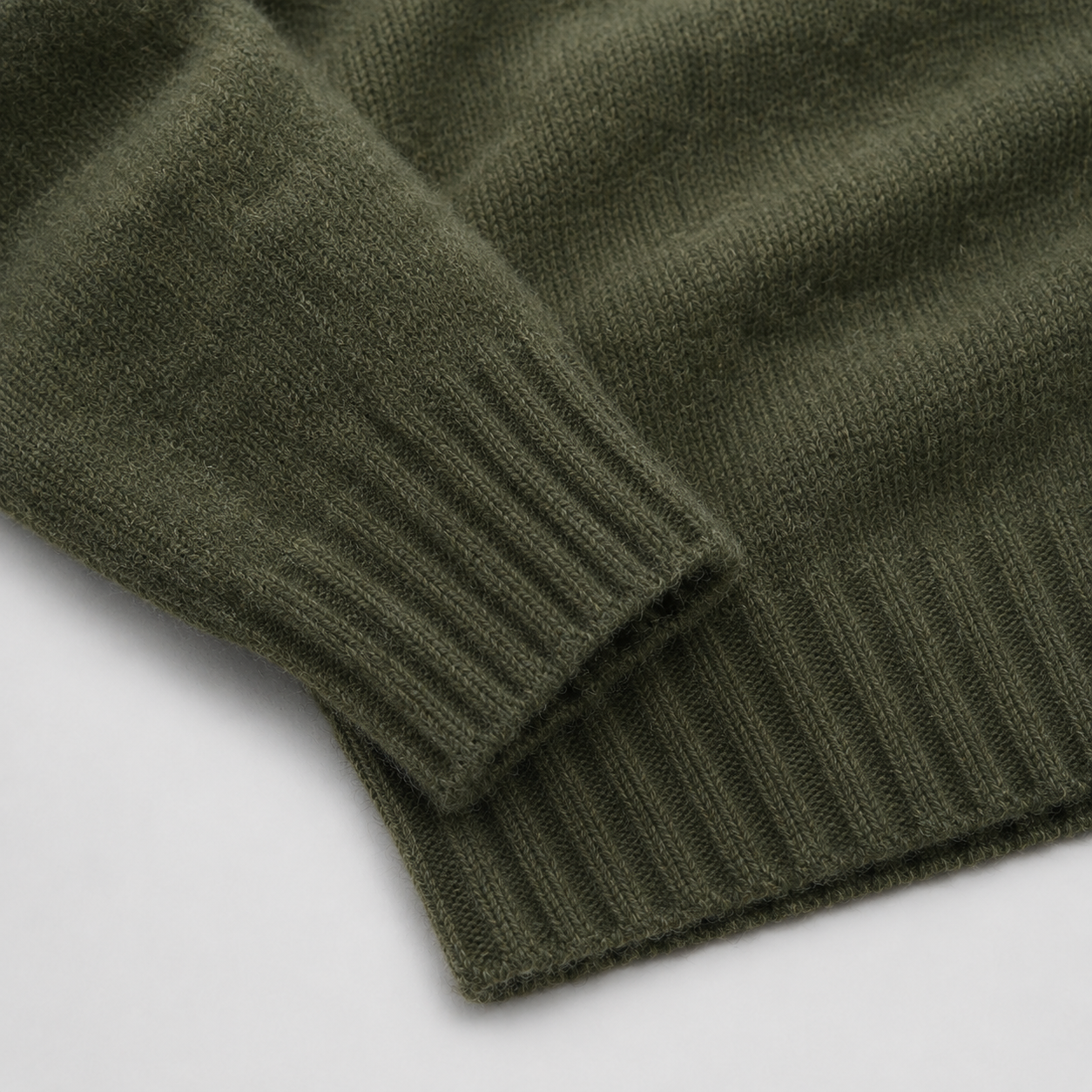 Close-up of an olive green knit sweater showing the ribbed cuff and hem detail against a light gray background.