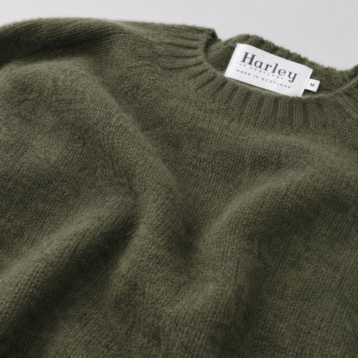 Close-up of a knit sweater in dark olive green, showing the ribbed crew neck and a white Harley brand label sewn inside.